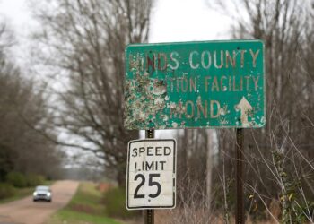A photo of a damaged green road sign that reads “Hinds County Detention Facility Raymond” with an arrow pointing forward.