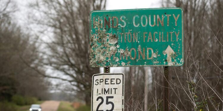 A photo of a damaged green road sign that reads “Hinds County Detention Facility Raymond” with an arrow pointing forward.
