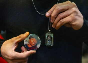 A photograph shows a closeup of Robert L. Brooks Jr., a Black man, holding a pin showing the face of his father — also a Black man — and also holding up a necklace with a pendant that has his father’s picture in it.