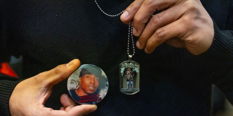 A photograph shows a closeup of Robert L. Brooks Jr., a Black man, holding a pin showing the face of his father — also a Black man — and also holding up a necklace with a pendant that has his father’s picture in it.