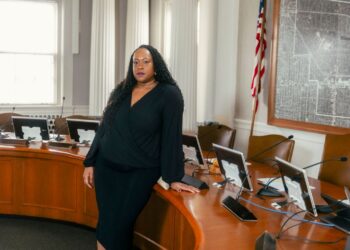 A Black woman with long black curly hair wearing a black dress stands in front of a brown desk used by the city council in Shaker Heights, Ohio. On the right, there is an American flag and a framed map on the wall.