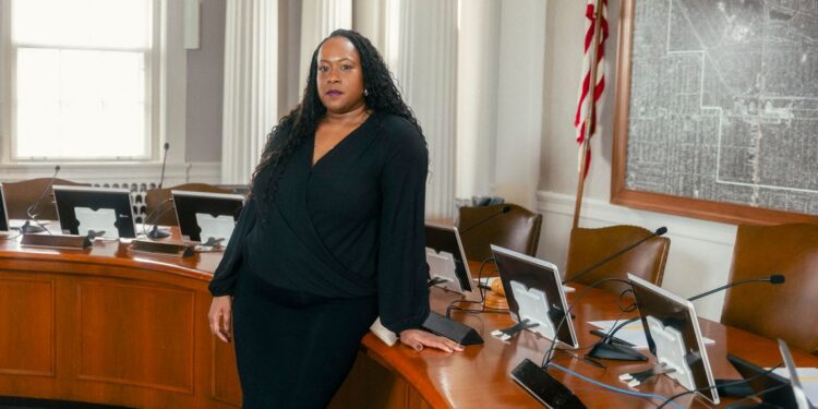 A Black woman with long black curly hair wearing a black dress stands in front of a brown desk used by the city council in Shaker Heights, Ohio. On the right, there is an American flag and a framed map on the wall.