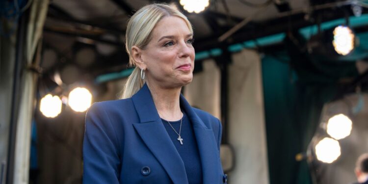 A woman with blonde hair wearing a blue suit walks in front of a tent with overhead lights.