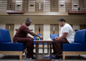 A person with dark-toned skin and a person with medium-toned skin, both wearing prison uniforms, play chess while sitting on blue chairs. In the background are doors to prison cells.