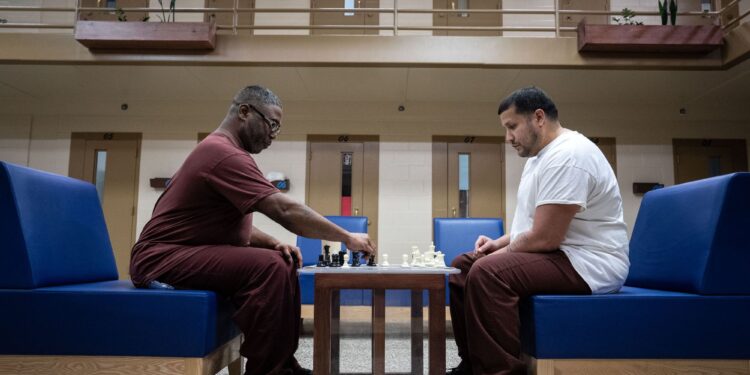 A person with dark-toned skin and a person with medium-toned skin, both wearing prison uniforms, play chess while sitting on blue chairs. In the background are doors to prison cells.