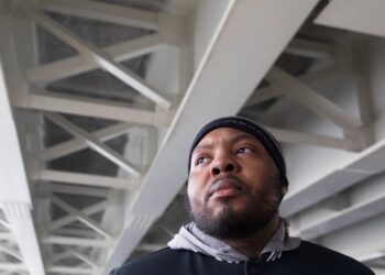 A photo of a Black man with a beard looking to the side under a concrete bridge. The man is wearing a black beanie and jacket and a gray hoodie.