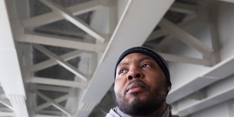 A photo of a Black man with a beard looking to the side under a concrete bridge. The man is wearing a black beanie and jacket and a gray hoodie.