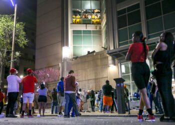 Outside a jail, a crowd watches people in light orange uniforms protesting by broken windows on a higher floor.