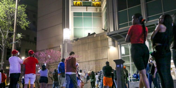 Outside a jail, a crowd watches people in light orange uniforms protesting by broken windows on a higher floor.