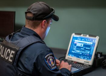 A police officer, wearing a cap, a vest and navy uniform, looks at a computer screen while sitting at desk.