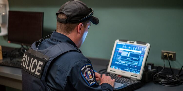 A police officer, wearing a cap, a vest and navy uniform, looks at a computer screen while sitting at desk.