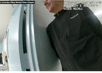 A man in a black zip-up jacket stands outside of a room while smiling.