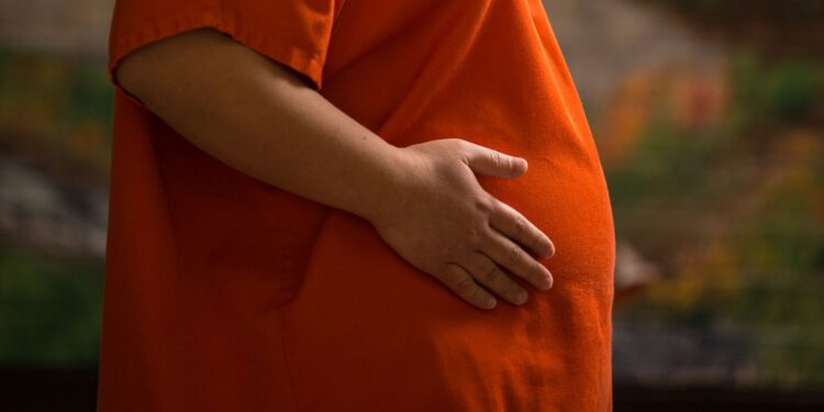 A pregnant woman wearing an orange prison uniform places her hand on her belly.