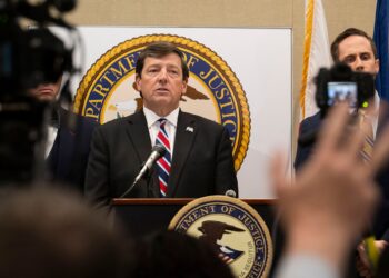 A White man, wearing a black suit and a blue and red tie, talks into a mic while standing at a podium with a U.S. Department of Justice seal on it. A group of reporters holding cameras and microphones stand in front of him.