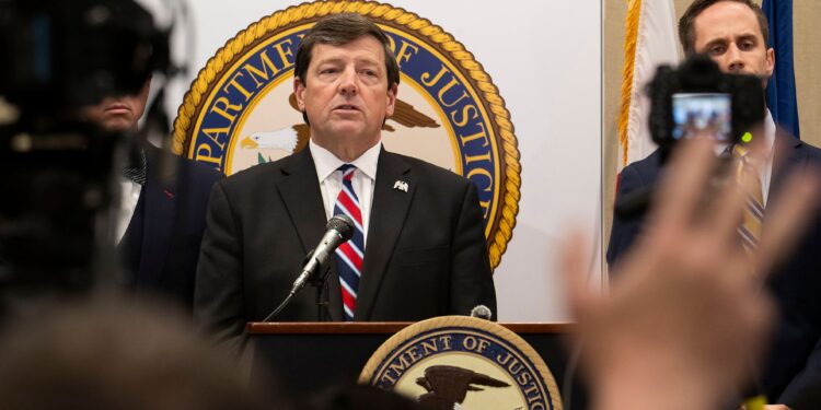 A White man, wearing a black suit and a blue and red tie, talks into a mic while standing at a podium with a U.S. Department of Justice seal on it. A group of reporters holding cameras and microphones stand in front of him.
