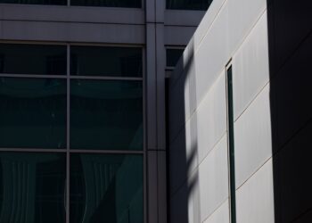 A photo shows a close view of the outside of a jail building. One side, in shade, has blue window panes. Another side, in harsh sunlight, shows a wall of concrete with a thin, vertical window.