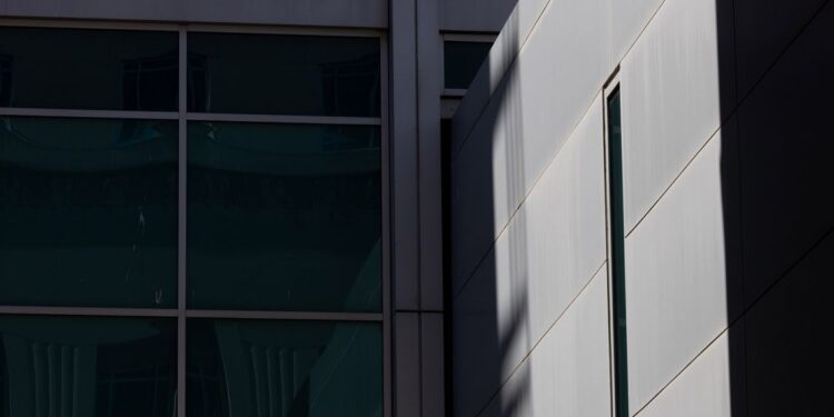 A photo shows a close view of the outside of a jail building. One side, in shade, has blue window panes. Another side, in harsh sunlight, shows a wall of concrete with a thin, vertical window.