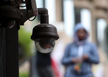 A photo shows a video surveillance camera in focus. A person in a light blue sweater is walking down a street in the background.