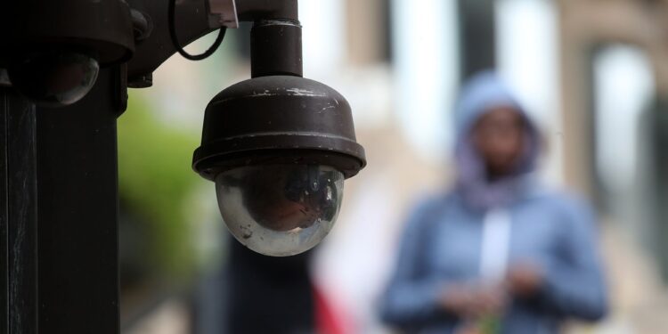 A photo shows a video surveillance camera in focus. A person in a light blue sweater is walking down a street in the background.