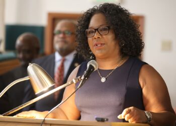 Louisiana Rep. Delisha Boyd, a Black woman wearing glasses and a black dress, speaks into a mic while standing in front of a podium. Two Black men wearing suits stand in the background.