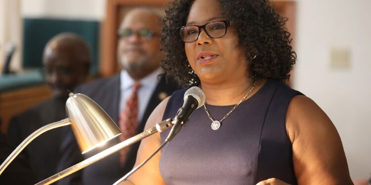Louisiana Rep. Delisha Boyd, a Black woman wearing glasses and a black dress, speaks into a mic while standing in front of a podium. Two Black men wearing suits stand in the background.