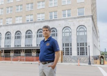 In a photograph, a White man with gray hair stands on the street in front of a concrete building with arched windows. The man is wearing a blue short-sleeved polo shirt with a logo.