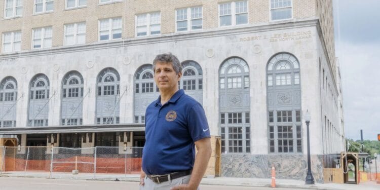 In a photograph, a White man with gray hair stands on the street in front of a concrete building with arched windows. The man is wearing a blue short-sleeved polo shirt with a logo.