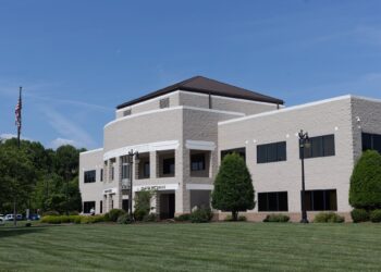 A photo of a beige, two-story building with a green lawn, under a blue sky with a few clouds.