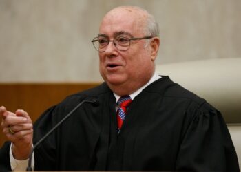 A photo of a White man behind a microphone gesturing while wearing a black judge’s robe.