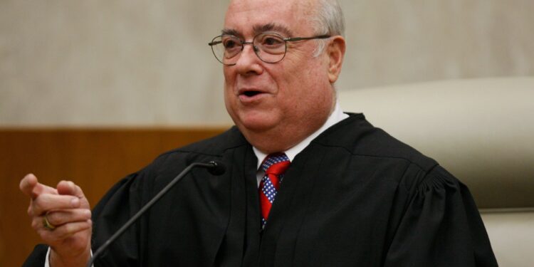 A photo of a White man behind a microphone gesturing while wearing a black judge’s robe.