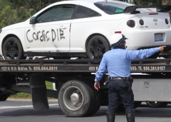 A white car with "Cordis DIE" spray-painted on its side being towed away by a tow truck, with a police officer overseeing.
