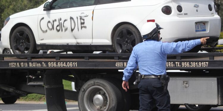 A white car with "Cordis DIE" spray-painted on its side being towed away by a tow truck, with a police officer overseeing.