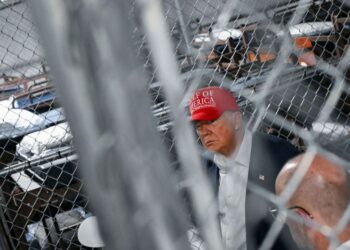 A photograph of a White man in his late 70s wearing a red hat with the words "Gulf of America" and a suit. He is seen through an out-of-focus chainlink fence and is looking to the side. Behind him there are rows of bunk beds behind another chainlink fence.
