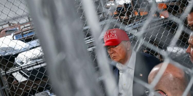 A photograph of a White man in his late 70s wearing a red hat with the words "Gulf of America" and a suit. He is seen through an out-of-focus chainlink fence and is looking to the side. Behind him there are rows of bunk beds behind another chainlink fence.