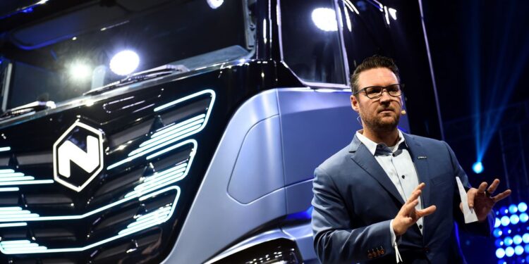 Trevor Milton, a man with light-toned skin, wearing glasses and a suit, stands indoors in front of a truck with blue and black panels.