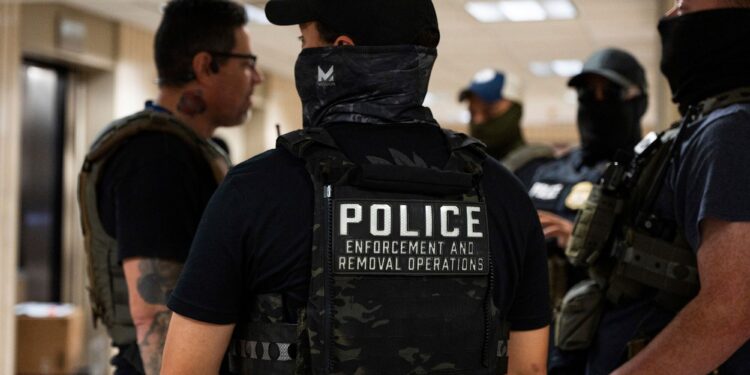 A photo shows the back of a light-skinned police officer wearing a black T-shirt, a black cap, face covering and a vest with the words "police enforcement and removal operations." There are 4 officers standing in the background.