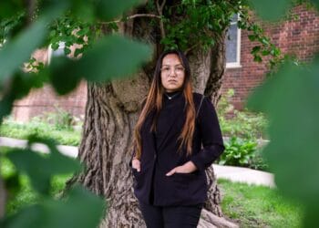A woman with light brown hair and glasses in a black suit jacket stands in front of a tree.