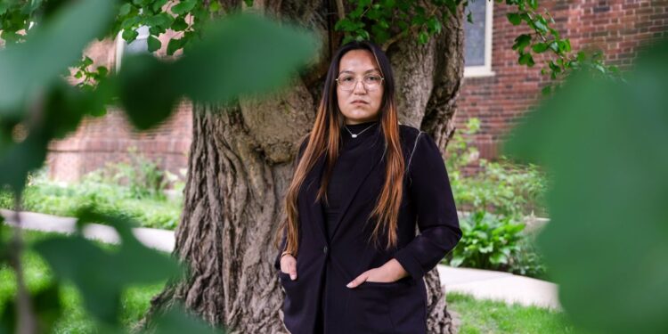 A woman with light brown hair and glasses in a black suit jacket stands in front of a tree.