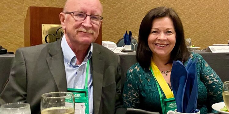 A elderly White man with a mustache, who is wearing glasses, a light blue button-down shirt, and a gray blazer, poses for a picture with his wife, a White woman with dark brown hair, who is wearing a green dress and a yellow ribbon.