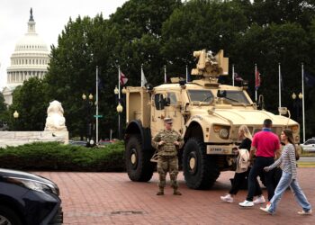 A person in a National Guard uniform stands next to a tan military vehicle parked on a red brick pavement. Three people walk in front of the military vehicle. The U.S. Capitol Dome and various trees can be seen in the background.