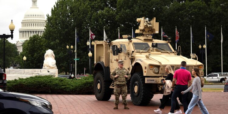 A person in a National Guard uniform stands next to a tan military vehicle parked on a red brick pavement. Three people walk in front of the military vehicle. The U.S. Capitol Dome and various trees can be seen in the background.