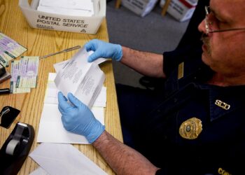A White male corrections officer wears light blue latex gloves as he reviews a letter on a wooden table. A United States Postal Service box with mail is on the table to his right.