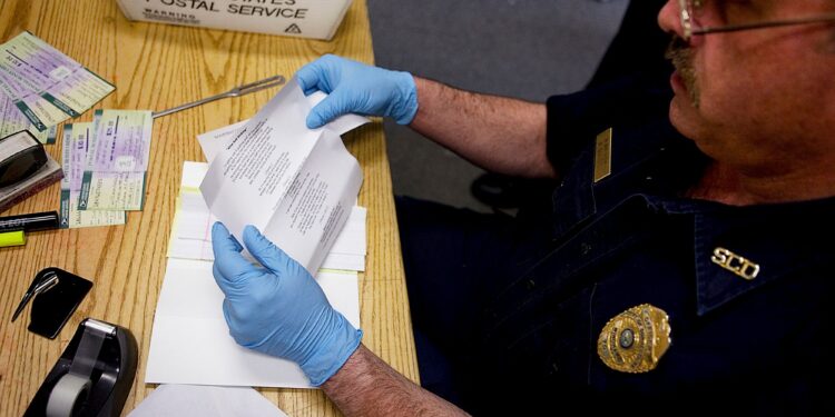 A White male corrections officer wears light blue latex gloves as he reviews a letter on a wooden table. A United States Postal Service box with mail is on the table to his right.