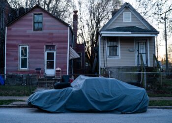 A photo shows a tarp-covered car parked on a street in front of a red house and a tan house.