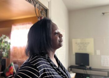 A Black woman wearing a black-and-white striped shirt stands in a room. A computer, a desk and a poster are in the background.