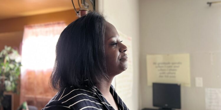 A Black woman wearing a black-and-white striped shirt stands in a room. A computer, a desk and a poster are in the background.