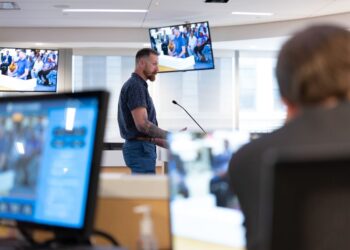 James DeCredico, a White man wearing a blue short-sleeved shirt and blue pants, stands at a podium in a county council meeting. There are two monitors showing the proceedings behind him.