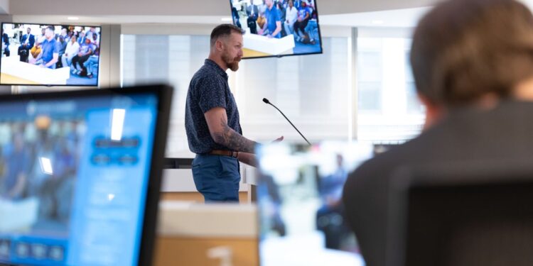 James DeCredico, a White man wearing a blue short-sleeved shirt and blue pants, stands at a podium in a county council meeting. There are two monitors showing the proceedings behind him.