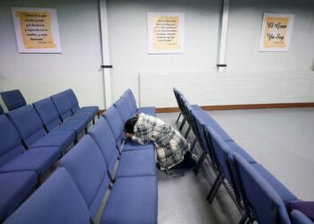A woman, wearing a black and white plaid jacket, kneels in between a row of blue chairs. Three signs of Bible verses translated into Spanish hang on the wall.