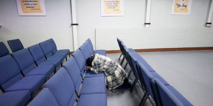 A woman, wearing a black and white plaid jacket, kneels in between a row of blue chairs. Three signs of Bible verses translated into Spanish hang on the wall.